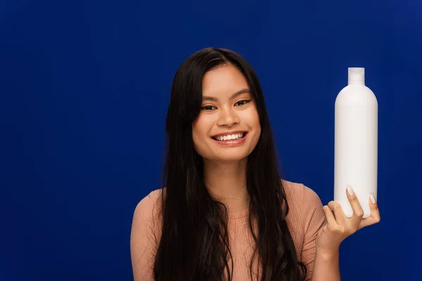 Smiling asian woman holding bottle of shampoo isolated on blue — Photo de stock