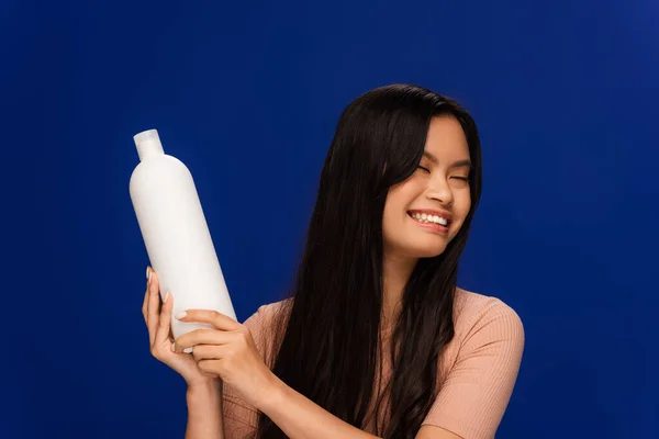 Cheerful asian woman holding bottle of shampoo isolated on blue — Fotografia de Stock