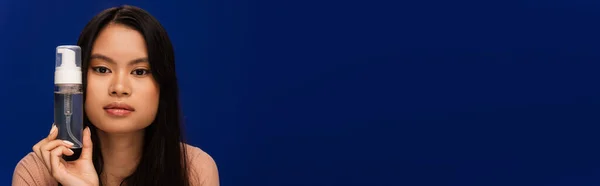 Young asian woman holding face foam and looking at camera isolated on blue, banner — Stock Photo