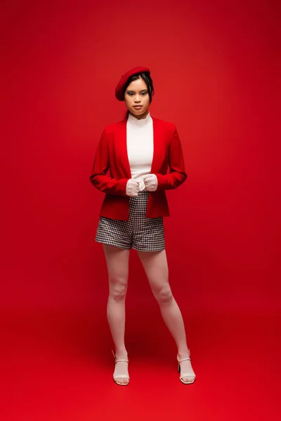 Full length of stylish asian model in beret looking at camera on red background — Stock Photo
