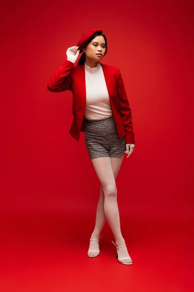 Asian woman in gloves touching beret and posing on red background — Stock Photo