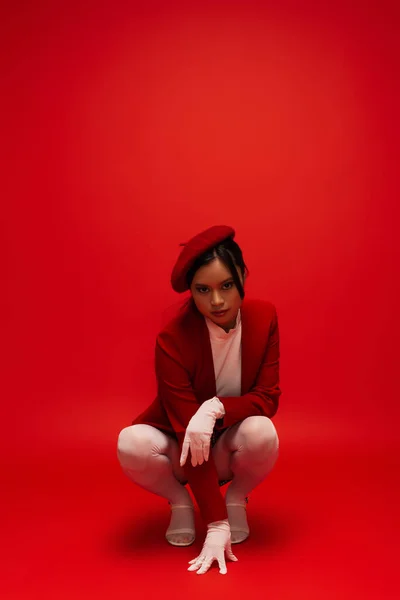 Fashionable asian woman in gloves and beret looking at camera on red background — Photo de stock