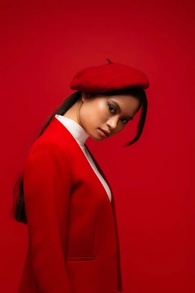 Портрет модної азіатської моделі в береті і блістері, дивлячись на камеру ізольовано на червоному — стокове фото