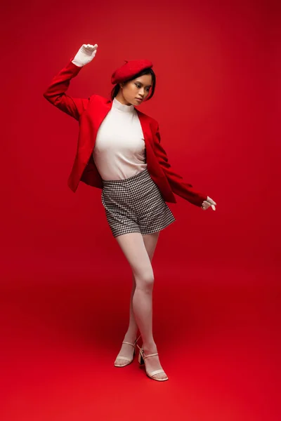 Full length of trendy asian woman in beret and blazer posing on red background — Foto stock
