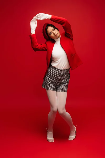 Full length of stylish asian model in gloves and beret posing with closed eyes on red background - foto de stock