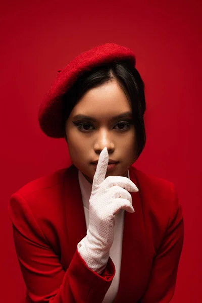 Portrait of asian model in beret showing secret gesture isolated on red — Stock Photo
