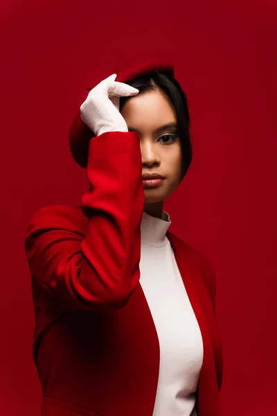 Trendy asian woman in jacket and glove adjusting beret isolated on red — Fotografia de Stock