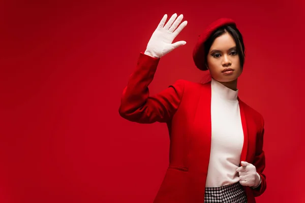 Asian woman in beret an gloves waving at camera isolated on red — Foto stock