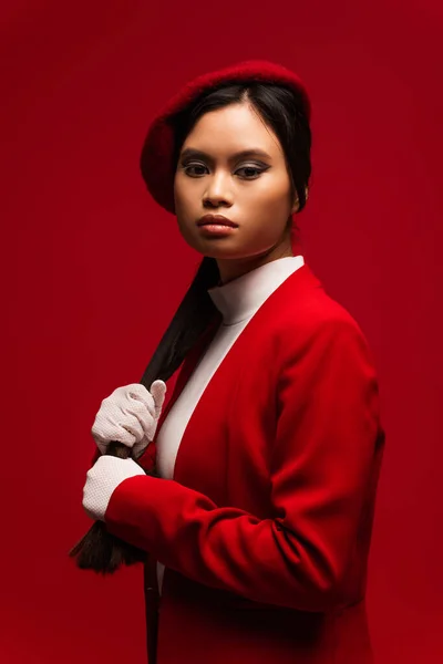 Brunette asian model in beret touching hair isolated on red — Stock Photo