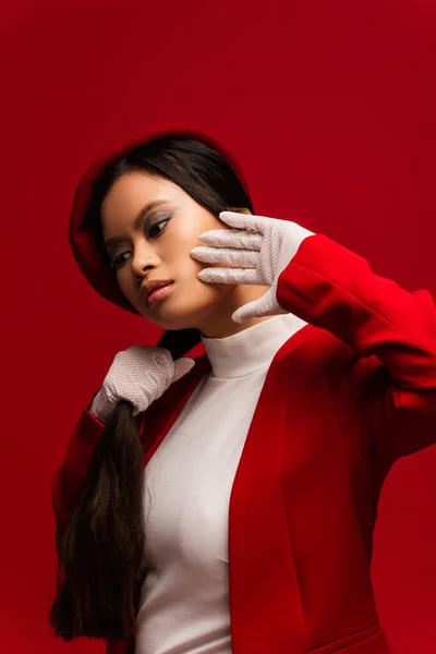 Fashionable asian model in jacket and gloves touching hair isolated on red - foto de stock