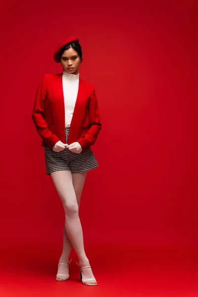 Full length of stylish asian woman in jacket and tights looking at camera on red background - foto de stock