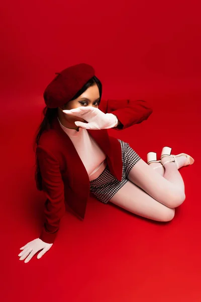 Overhead view of trendy asian model in white coves covering face while sitting on red background — Stock Photo