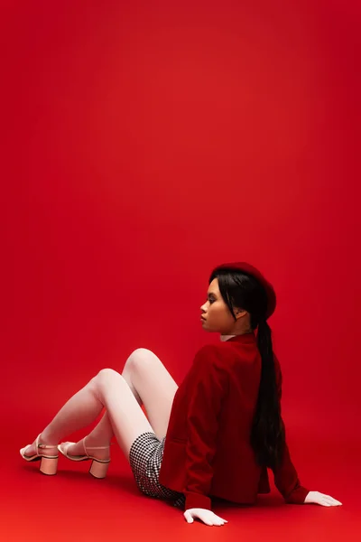 Side view of stylish asian model in white tights and gloves looking away while sitting on red background — Stock Photo