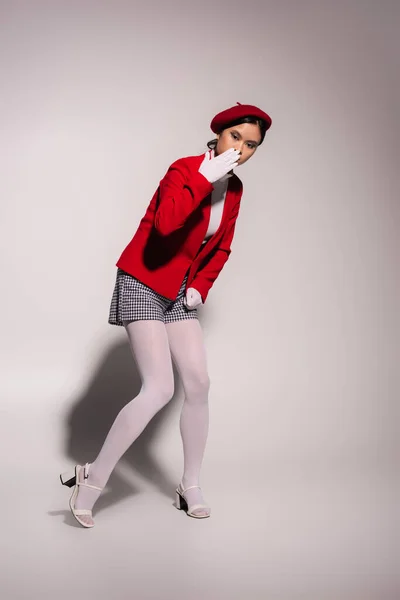 Stylish asian woman in red beret and gloves holding hand near mouth on grey background — Photo de stock