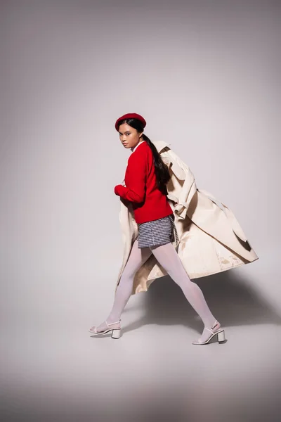 Full length of fashionable asian woman in red beret holding trench coat while walking on grey background — Foto stock