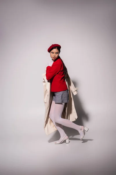 Full length of stylish asian woman in red jacket and beret holding trench coat on grey background — Stockfoto