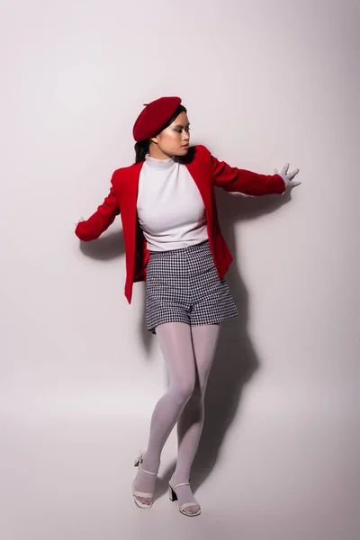 Full length of trendy asian model in red beret and jacket touching grey background — Foto stock
