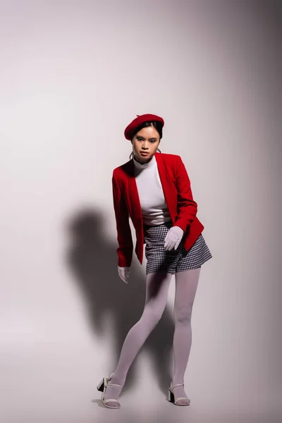 Stylish asian woman in red beret and white tights looking at camera on grey background with shadow — Foto stock