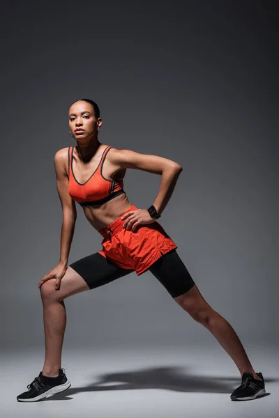 Повна довжина молодої афроамериканської жінки в спортивному одязі і кросівки, вправи з рукою на стегні на сірому — стокове фото