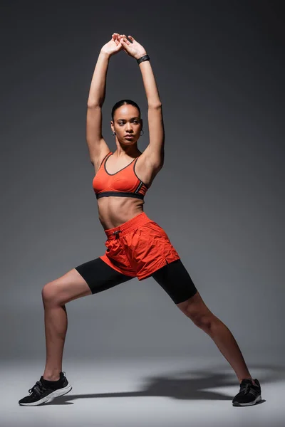 Full length of young african american sportswoman in sneakers exercising with raised hands on grey — Fotografia de Stock
