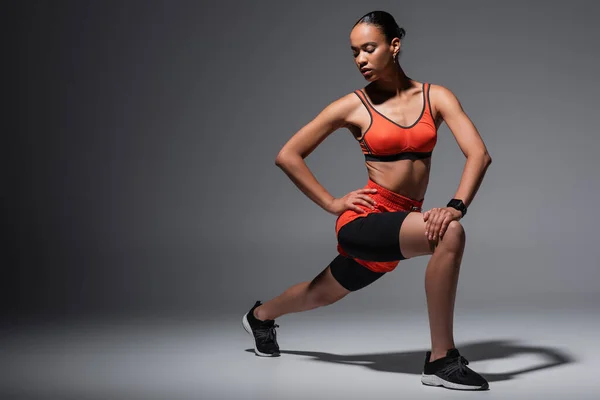 Повна довжина молодої афроамериканської жінки в спортивному одязі і кросівки, вправи з рукою на стегні на сірому — стокове фото