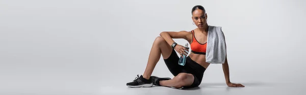 Young african american woman in sports bra and bike shorts sitting with sports bottle and towel on grey background, banner — стоковое фото
