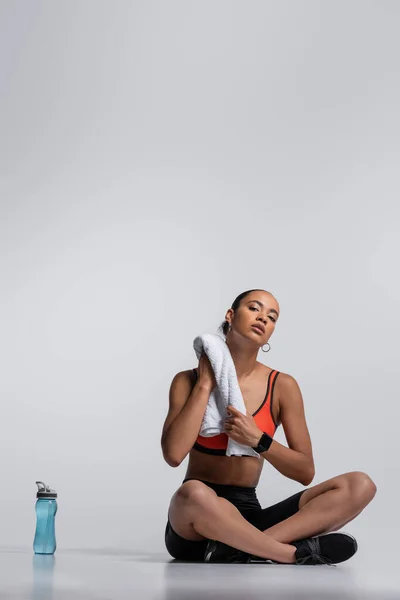 Full length of brunette african american sportswoman sitting with towel near sports bottle on grey — Stock Photo