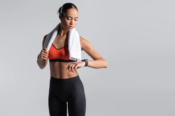Brunette african american sportswoman in red sports bra standing with towel and looking at fitness tracker isolated on grey — Fotografia de Stock