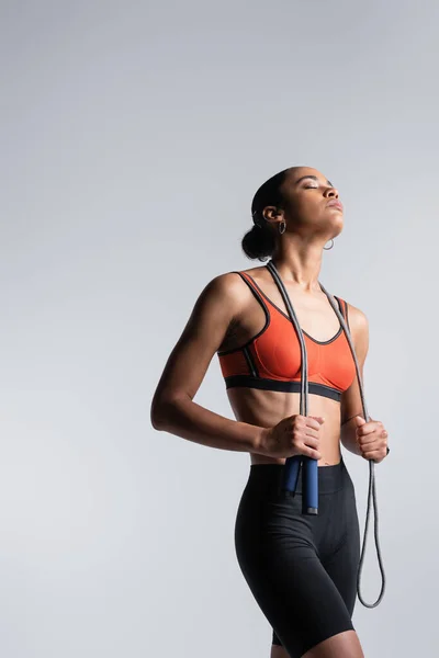 Young african american sportswoman with closed eyes holding skipping rope isolated on grey — Stockfoto