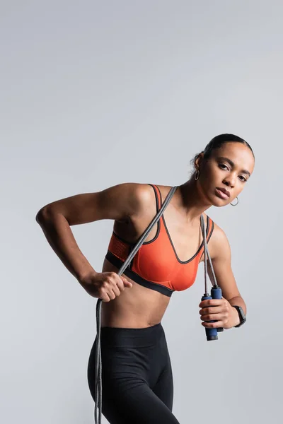 Young african american sportswoman in red sports bra and bike shorts holding skipping rope isolated on grey — Stock Photo