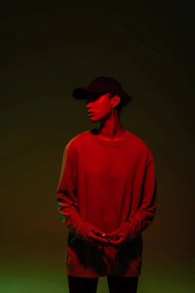 Young african american woman in baseball cap and sweatshirt standing on green with red light — Stockfoto