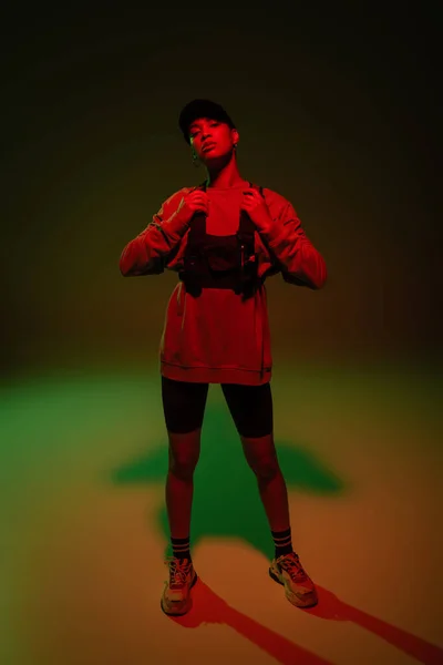 Full length of young african american woman in baseball cap and sweatshirt adjusting vest while standing on green - foto de stock