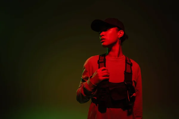 Young african american woman in baseball cap and sweatshirt standing on green background with red light — Stockfoto