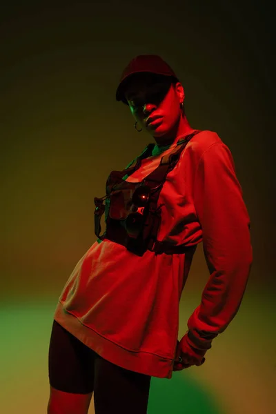 African american woman in baseball cap and sweatshirt standing on green with red light - foto de stock