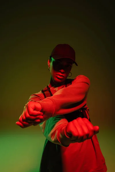 African american woman in sweatshirt and baseball cap posing on green with red light — Stockfoto