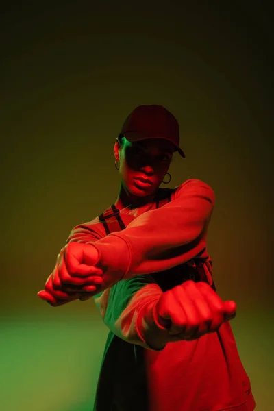 African american woman in sweatshirt and baseball cap posing on green with red light — стоковое фото