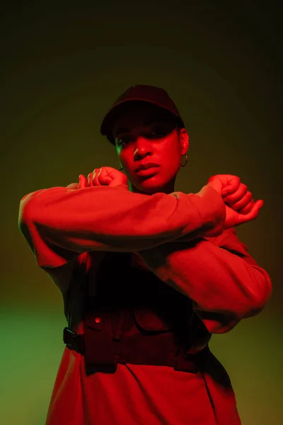 African american woman in sweatshirt and baseball cap posing with crossed arms on green with red light — Stockfoto
