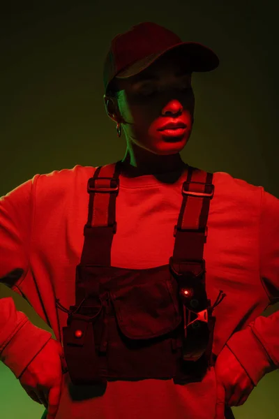 African american woman in sweatshirt and vest posing with hands on hips on green with red light - foto de stock