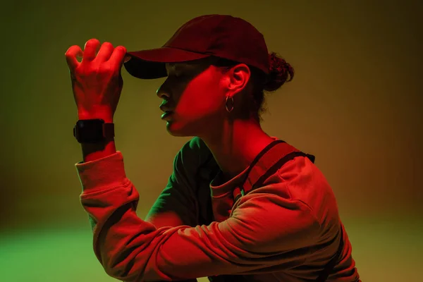 Side view of african american woman in sweatshirt adjusting baseball cap on green with red light - foto de stock