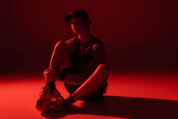 Full length of young african american woman in baseball cap and sweatshirt sitting on dark red background — стоковое фото