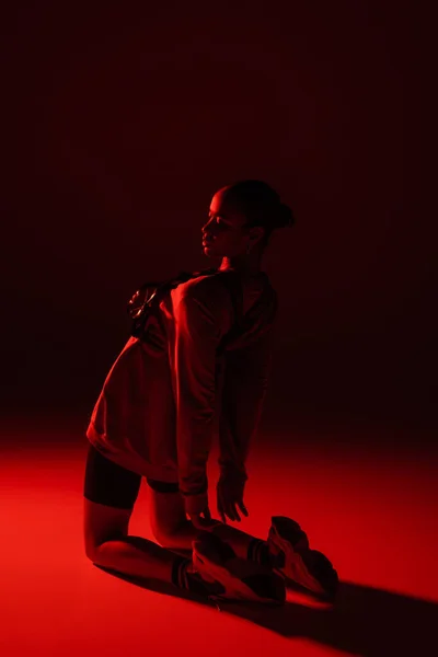 Full length of young african american woman in bike shorts and sweatshirt posing on dark red background - foto de stock