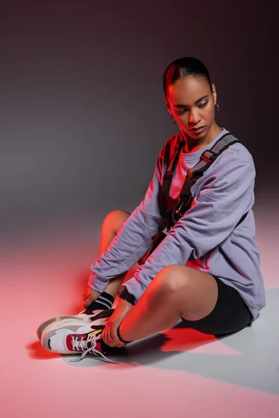 Full length of stylish african american woman in bike shorts and sweatshirt sitting on grey with red light — стоковое фото