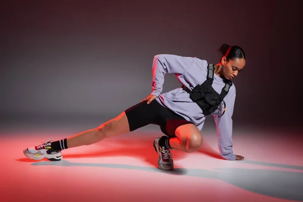 Full length of african american woman in bike shorts and sweatshirt stretching leg on grey background with red light — Stock Photo