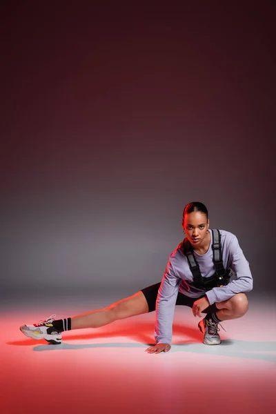 Full length of sporty african american woman in bike shorts and sweatshirt stretching leg on grey with red light — Stock Photo