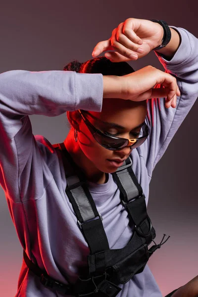 Portrait of african american woman in sweatshirt and trendy sunglasses looking down while posing on grey with red light — Stock Photo
