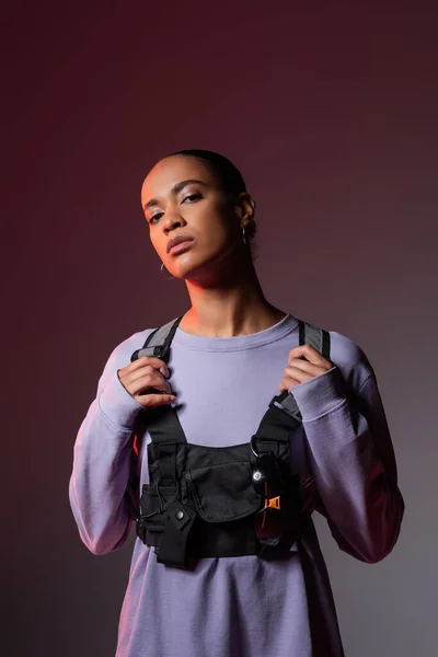 Young african american woman in sweatshirt and vest looking at camera on grey and purple — Stock Photo