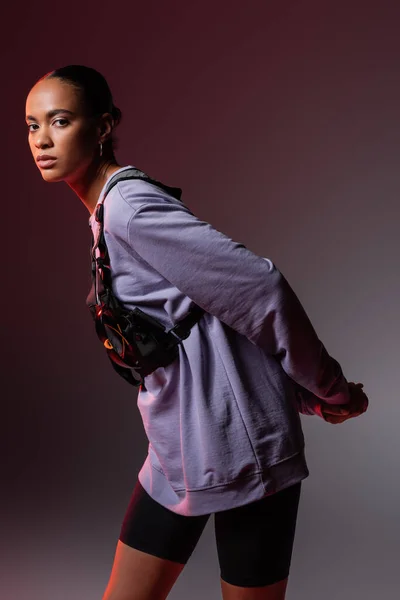 Young african american woman in sweatshirt and vest looking at camera on grey and purple — Stock Photo