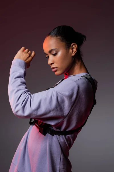 Brunette african american woman in sweatshirt and vest posing on grey and purple — Stock Photo