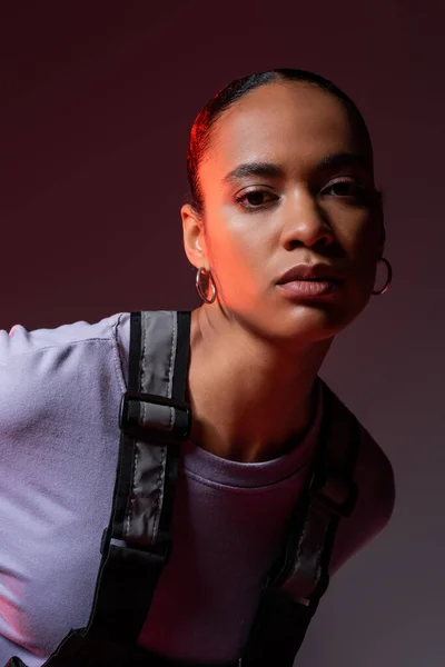 Brunette african american woman in sweatshirt and vest looking at camera on dark purple — Stock Photo