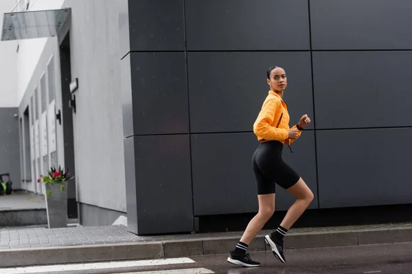 Full length of brunette african american sportswoman in bike shorts and yellow puffer jacket running outside - foto de stock